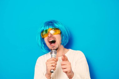 Peruklu ve güneş gözlüklü bir adam mikrofona şarkı söylüyor..