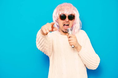 Peruklu ve güneş gözlüklü bir adam mikrofona şarkı söylüyor..