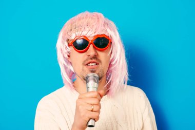 Peruklu ve güneş gözlüklü bir adam mikrofona şarkı söylüyor..