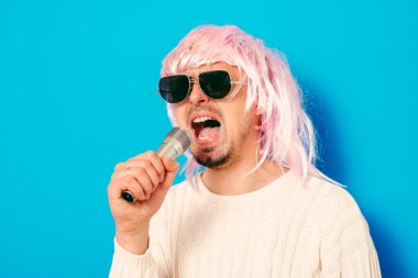 Peruklu ve güneş gözlüklü bir adam mikrofona şarkı söylüyor..