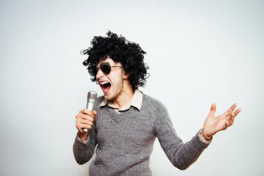 Peruklu ve güneş gözlüklü bir adam mikrofona şarkı söylüyor..