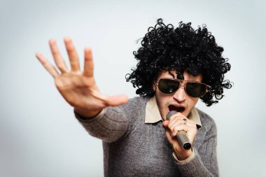 Peruklu ve güneş gözlüklü bir adam mikrofona şarkı söylüyor..