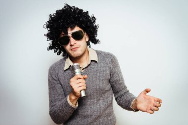 Peruklu ve güneş gözlüklü bir adam mikrofona şarkı söylüyor..