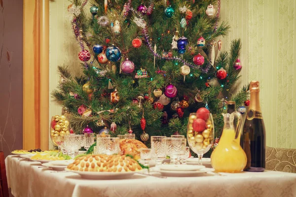 Mesa servida con platos festivos cerca de hermoso árbol de Navidad decorado en el interior del salón. Concepto de vacaciones de año nuevo en casa acogedora. Céntrate en el abeto, tono vintage — Foto de Stock