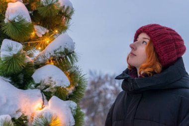 Kışın City caddesinde süslü Noel ağacının yanında duran güzel bir bayanın portresi. Genç kız akşamları yeni yıl ladin 'i arıyor. Şehir tatili, kış tatili, şehir konseptinde tatiller.