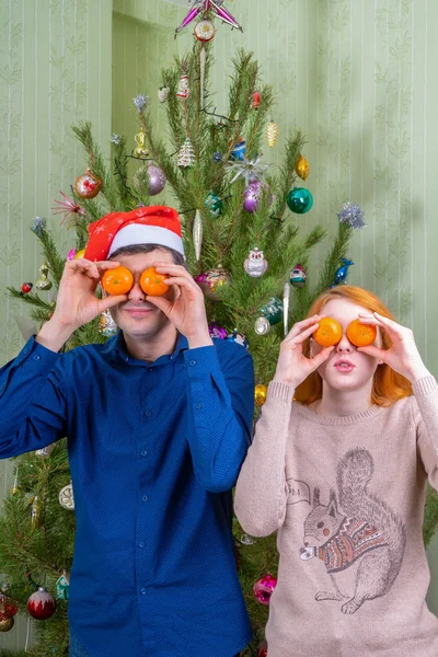 Hombre de mediana edad y hermosa joven divirtiéndose contra el árbol de año nuevo decorado. Padre e hija celebrando la Navidad con naranjas mandarinas. Vacaciones de invierno en el acogedor concepto de casa —  Fotos de Stock