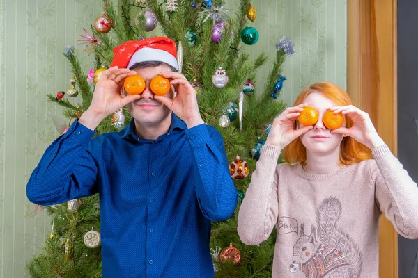 Hombre de mediana edad y hermosa joven divirtiéndose contra el árbol de año nuevo decorado. Padre e hija celebrando la Navidad con naranjas mandarinas. Vacaciones de invierno en el acogedor concepto de casa —  Fotos de Stock