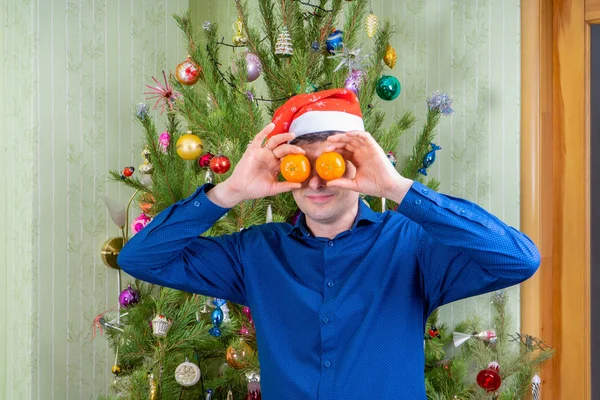 Guapo hombre de mediana edad en el sombrero de Santa Claus divertirse contra hermoso árbol de año nuevo decorado. Hombre celebrando la Navidad con mandarinas naranjas. Vacaciones de invierno en el acogedor concepto de casa —  Fotos de Stock