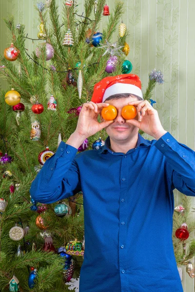 Guapo hombre de mediana edad en el sombrero de Santa Claus divertirse contra hermoso árbol de año nuevo decorado. Hombre celebrando la Navidad con mandarinas naranjas. Vacaciones de invierno en el acogedor concepto de casa —  Fotos de Stock