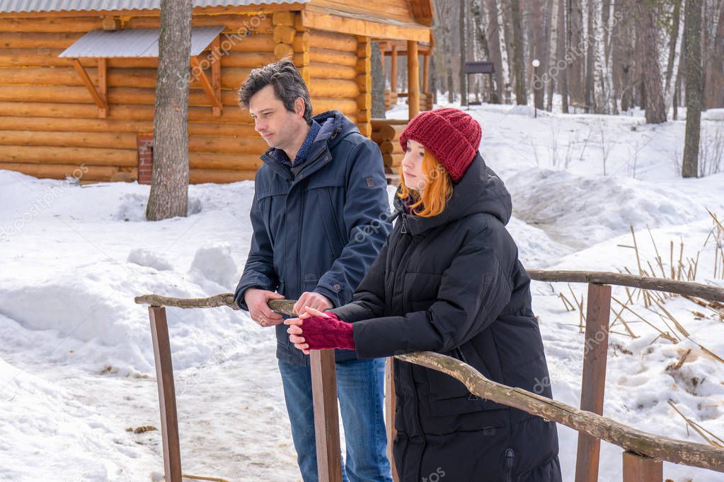 Father and daughter spending weekend at countryside in winter. Handsome middle-aged man and beautiful young lady standing on wooden bridge against country house and forest. People outdoors