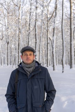 Karlı parkta ya da ormanda yürüyen orta yaşlı yakışıklı bir adamın portresi. Ceketli, atkılı ve şapkalı çekici bir adam kameraya bakıyor. Kış havası, otantik yaşam tarzı, şık erkek kıyafeti.