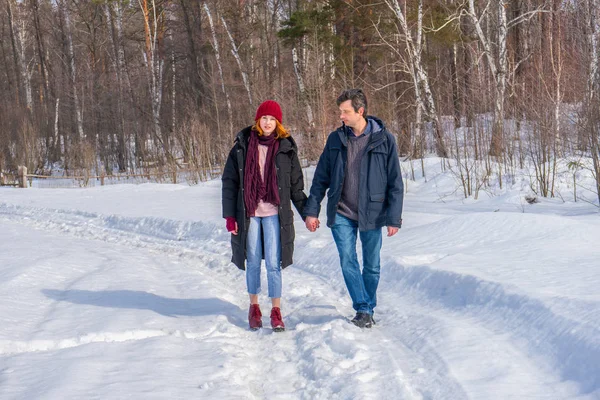 Handsome man and attractive young woman walking along snowy country road in sunny day. Beautiful look, male and female fashion, winter outfit. Winter holidays, weekend at countryside concept