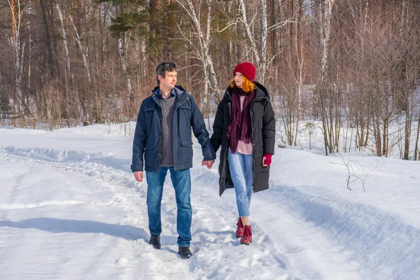 Handsome man and attractive young woman walking along snowy country road in sunny day. Beautiful look, male and female fashion, winter outfit. Winter holidays, weekend at countryside concept