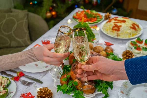 Köpüklü şarapla bardakları tutan insan elleri. Arkadaşlar ya da aile şenlikli Noel masasına ve süslü yılbaşı ağacına şampanyayla kadeh kaldırıyor. Konforlu evde kış tatili kutlaması — Stok fotoğraf
