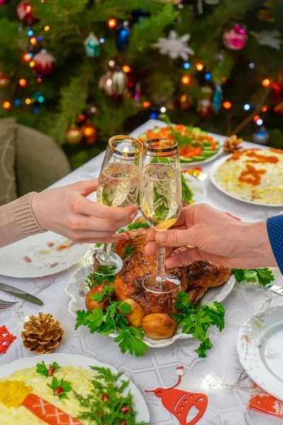 Köpüklü şarapla bardakları tutan insan elleri. Arkadaşlar ya da aile şenlikli Noel masasına ve süslü yılbaşı ağacına şampanyayla kadeh kaldırıyor. Konforlu evde kış tatili kutlaması — Stok fotoğraf