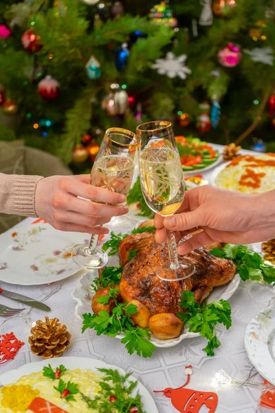 Köpüklü şarapla bardakları tutan insan elleri. Arkadaşlar ya da aile şenlikli Noel masasına ve süslü yılbaşı ağacına şampanyayla kadeh kaldırıyor. Konforlu evde kış tatili kutlaması — Stok fotoğraf