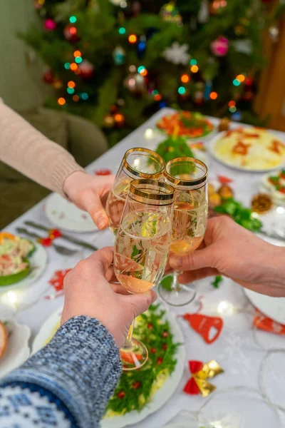 Menschenhände halten Gläser mit Sekt in der Hand. Freunde oder Familie stoßen mit Champagner vor festlicher Weihnachtstafel und geschmücktem Neujahrsbaum an. Winterferienfeier im gemütlichen Heim — Stockfoto