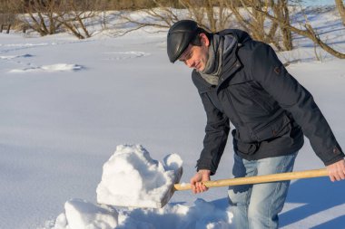 Güneşli bir günde ev sahasında kar temizleyen yakışıklı orta yaşlı beyaz bir adam. Kır evinden yola kürek döşeyen bir adam. Kış aktif eğlence kavramı