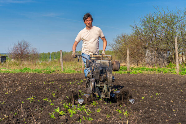 Man in wellingtons with cultivator ploughing ground in sunny day. Farmer plowing kitchen-garden in suburb. Land cultivation, soil tillage. Spring work in garden. Gardening concept.