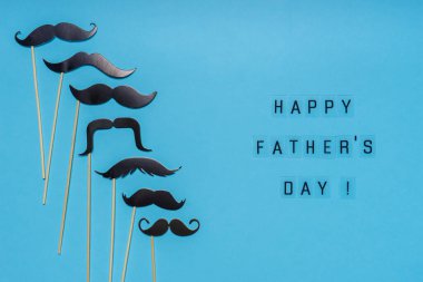 Çeşitli siyah fotoğraf kulübeleri mavi arka planda farklı şekildeki bıyıkları destekliyor. Tebrik kartı, mutlu babalar günü mesajı. Düz yatış, üst görünüm.