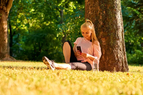 Young woman sitting at a tree in a park with her phone — Stock Photo, Image