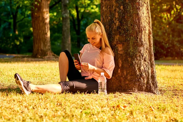 Young woman sitting at a tree in a park with her phone — Stock Photo, Image