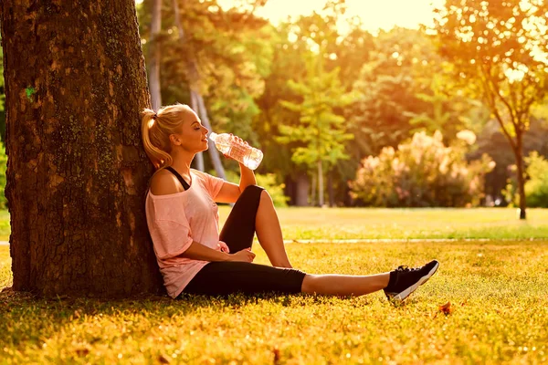 Giovane donna seduta ad un albero in un parco e acqua potabile — Foto Stock