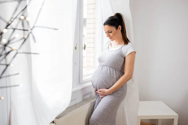 Een zwangere vrouw die naast het raam staat met handen op haar b — Stockfoto