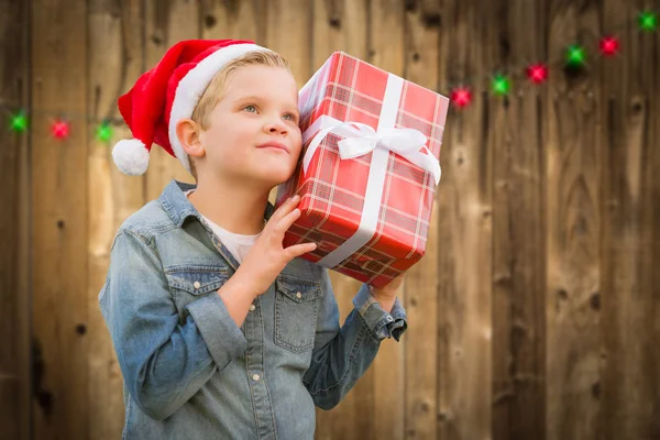 Garoto curioso vestindo chapéu de Santa segurando presente de Natal na madeira — Fotografia de Stock