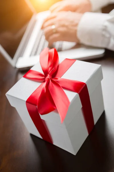 Λευκό δώρο κουτί με κόκκινη κορδέλα και φιόγκο κοντά άνθρωπος πληκτρολογώντας σε φορητό υπολογιστή — Φωτογραφία Αρχείου
