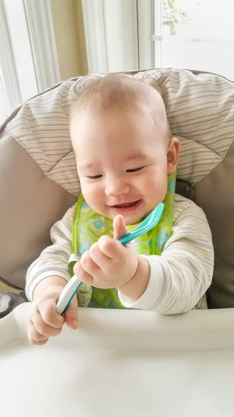 Adorable niño chino y caucásico jugando con su cuchara sentada en una silla alta . — Foto de Stock