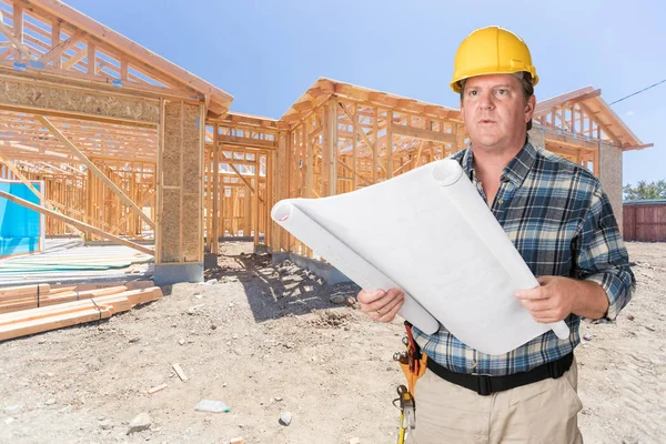 Male Contractor With House Plans Wearing Hard Hat In Front of New House Construction Framing