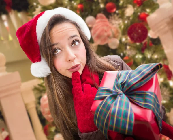 Ragazza pensante che indossa un cappello di Natale Babbo Natale con fiocco avvolto regalo di fronte all'albero decorato . — Foto Stock