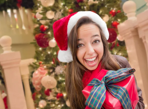 Ragazza che indossa un cappello di Babbo Natale con fiocco avvolto regalo di fronte all'albero decorato — Foto Stock