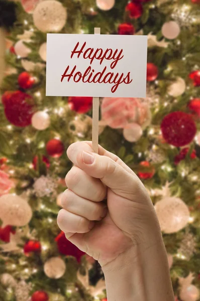 Χέρι που κρατάει την κάρτα Happy Holidays μπροστά από το στολισμένο χριστουγεννιάτικο δέντρο. — Φωτογραφία Αρχείου