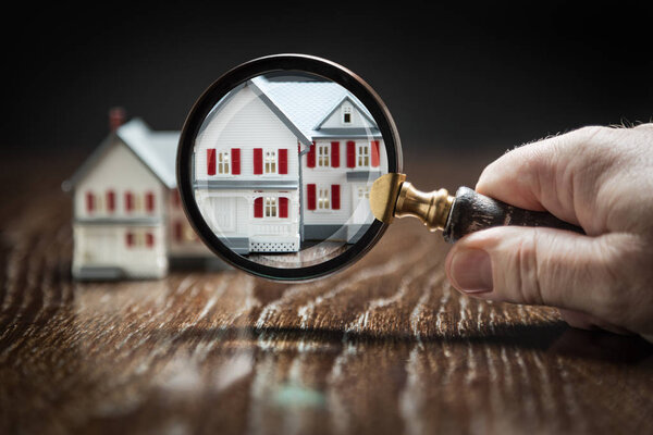 Hand Holding Magnifying Glass Up To Model Home on Reflective Wooden Surface.