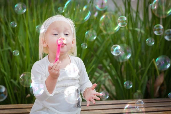 Schattig meisje, zittend op de Bank met het blazen van zeepbellen buiten plezier. — Stockfoto