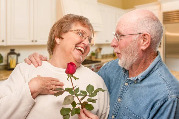 Feliz homem adulto sênior dando rosa vermelha para sua esposa dentro da cozinha . — Fotografia de Stock