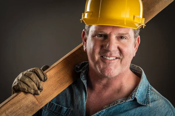 Lächelnder Bauunternehmer mit hartem Hut, der Holzplanke mit dramatischer Beleuchtung hält. — Stockfoto