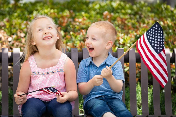 Hermana joven y hermano comparándose tamaño de la bandera americana en el banco en el parque . — Foto de Stock