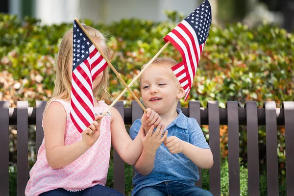 Kleine Schwester und Bruder schwenken amerikanische Flaggen auf der Parkbank. — Stockfoto