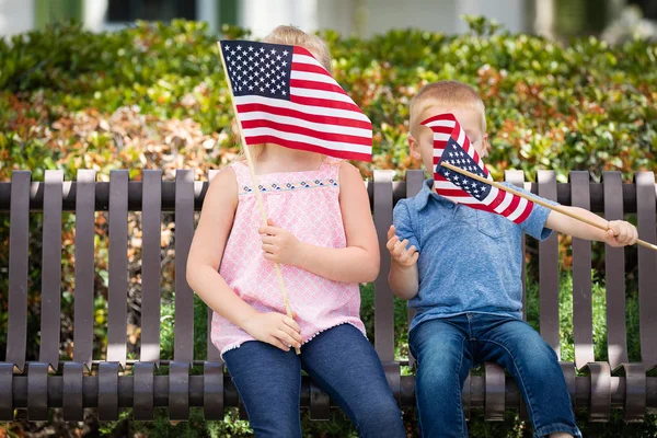 Kleine Schwester und Bruder schwenken amerikanische Flaggen auf der Parkbank. — Stockfoto