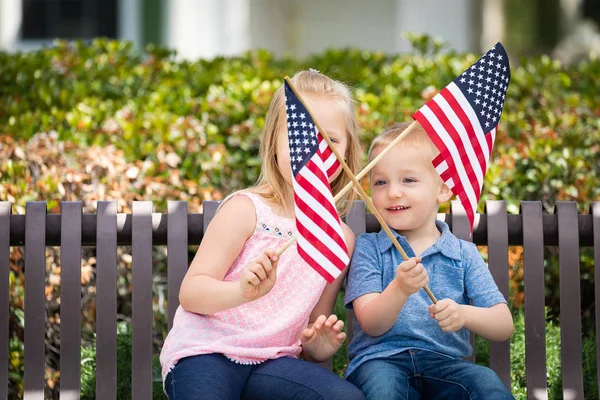 Молодая сестра и брат размахивают американскими флагами на скамейке в парке . — стоковое фото