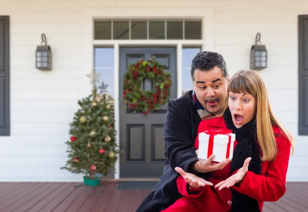 Pareja joven de raza mixta intercambiando regalos en el porche delantero de la casa con decoraciones navideñas . — Foto de Stock