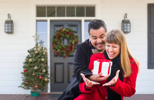 Young Mixed Race Couple Exchanging Gift On Front Porch of House with Christmas Decorations. — Stock Photo, Image