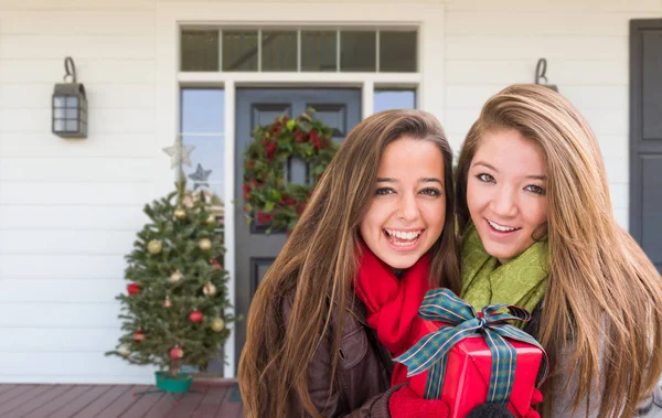 Jóvenes niñas de raza mixta sosteniendo regalo envuelto de pie en la Navidad decorado porche delantero —  Fotos de Stock