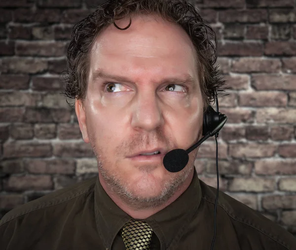 Empresario confuso poco amigable con cara fruncida que usa auriculares para teléfono en la oficina con fondo de ladrillo —  Fotos de Stock