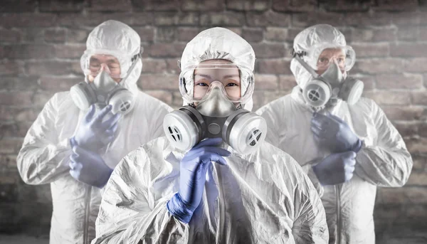 Mujer china y equipo detrás en suites de materiales peligrosos, máscaras de gas y gafas contra la pared de ladrillo —  Fotos de Stock