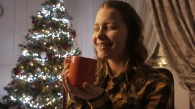 Genç kız gülümseyerek ve bir fincan çay içme. Noel ağacı ve tatil ruh hali. 4k Uhd.