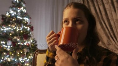 Genç kız gülümseyerek ve bir fincan çay içme. Noel ağacı ve tatil ruh hali. 4k Uhd.